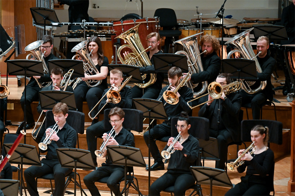 Students performing a variety of brass students, seated in a brass section to the right of the conductor, wearing formal attire.